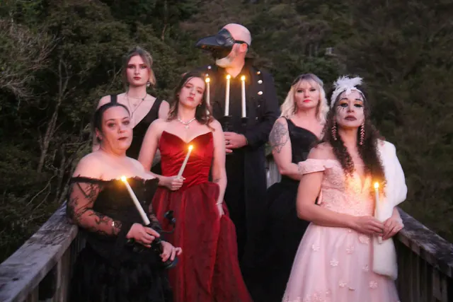 Six guests in ball gowns holding lit candles on a wooden bush bridge at dusk, a figure in a plague doctor mask and long coat standing behind them with a candelabra