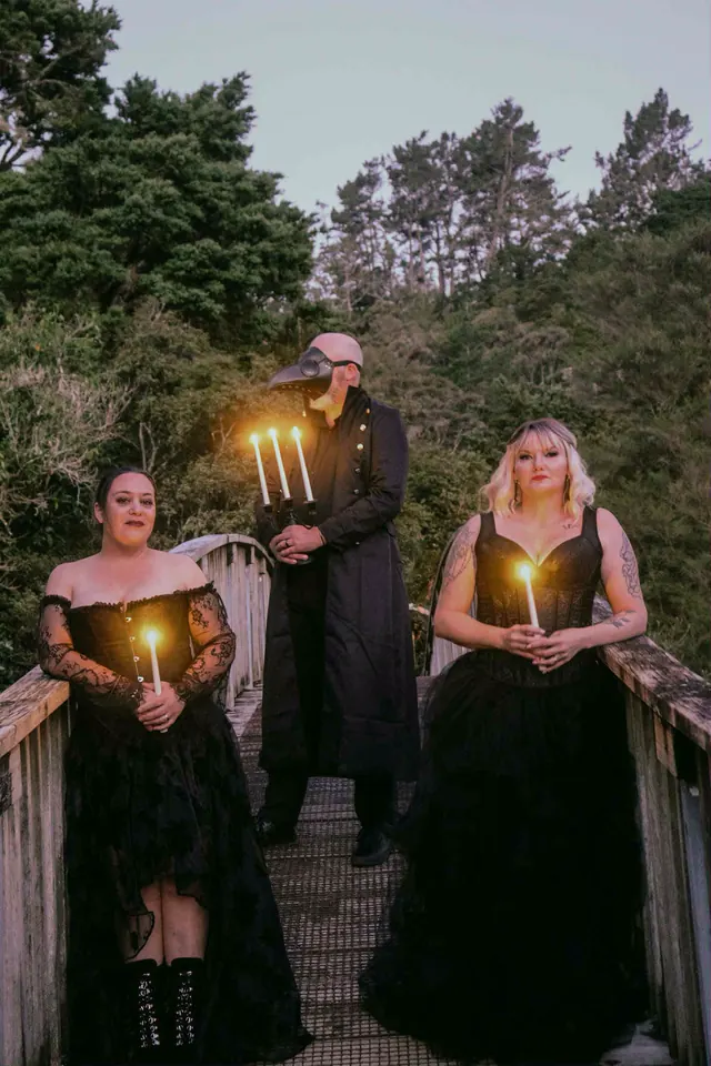 Two women in black lace ball gowns holding candles at either end of a bush footbridge, the plague doctor between them with a three-branch candelabra
