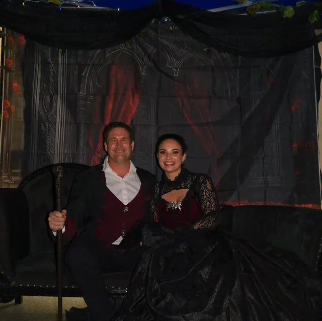 A couple in Victorian evening dress seated in the gothic photo booth, him with a cane and a wine-red waistcoat, her in black lace over deep red