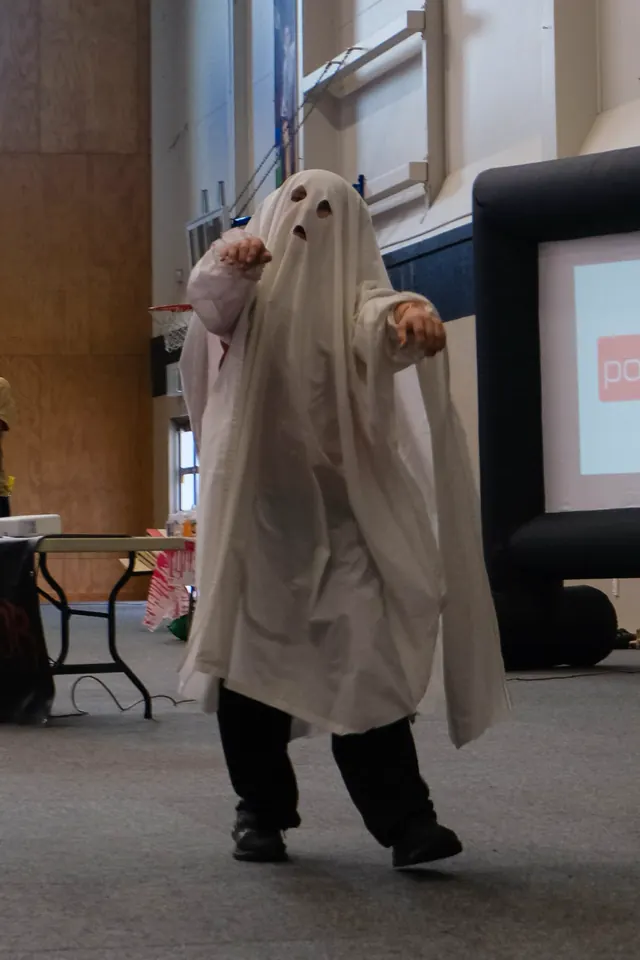 A child in a bedsheet ghost costume with cut-out eye holes walking arms-out across the hall at a trick or treat day
