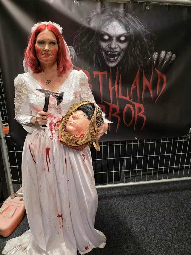 A blood-spattered bride holding a hatchet and a severed rubber head in a string basket, standing in front of the Northland Horror banner