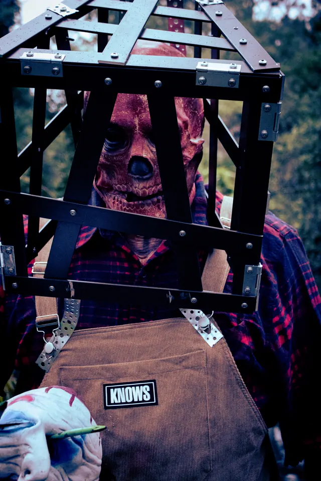 Close-up of the caged scarecrow - a raw, skinned-looking face pressed against the bars of a steel frame worn as a helmet