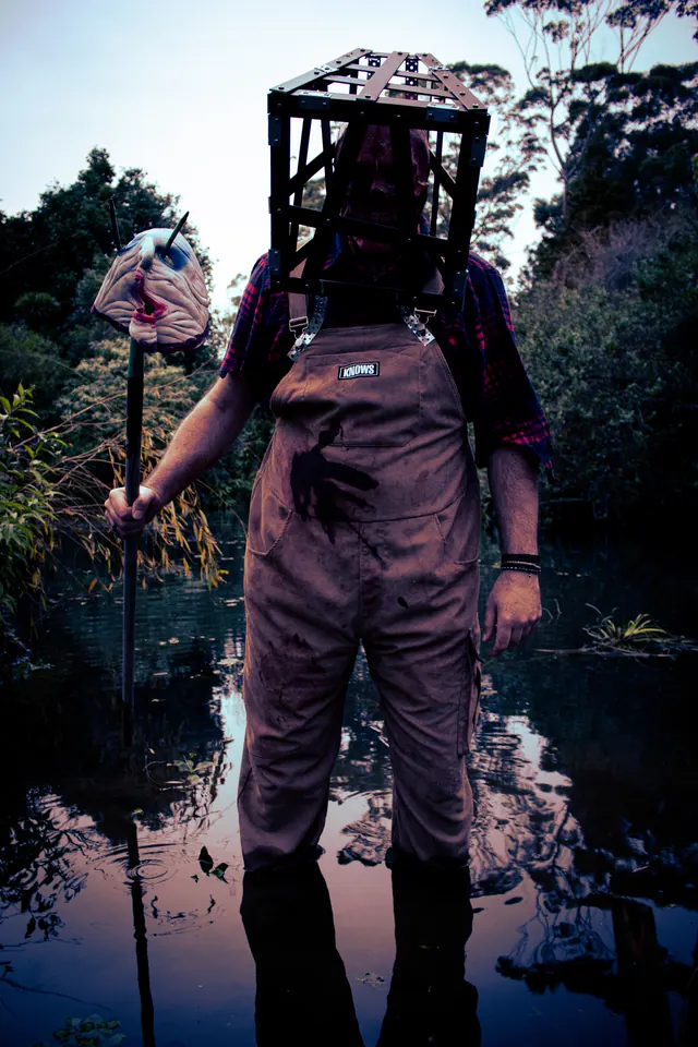 A figure in muddy overalls stands ankle-deep in still water at dusk with a steel cage locked over its head, holding a pole topped with a severed rubber head