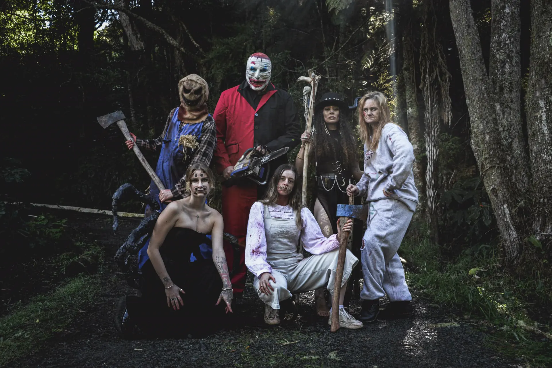 Six Northland Horror performers grouped on a bush track at dusk, a scarecrow in a burlap hood with an axe, a clown in a red and black suit holding a chainsaw, and a horned figure with a skull-topped staff
