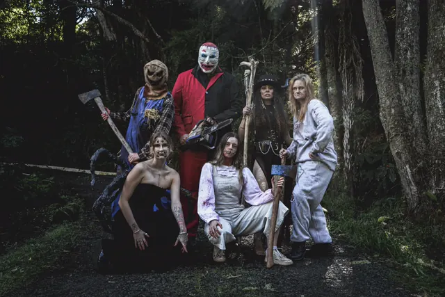 Six Northland Horror performers grouped on a bush track at dusk, a scarecrow in a burlap hood with an axe, a clown in a red and black suit holding a chainsaw, and a horned figure with a skull-topped staff
