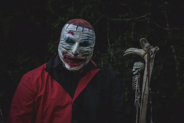 Close-up of a clown mask with a scalped red crown and a wide stitched grin, worn with a red and black suit, against dark bush