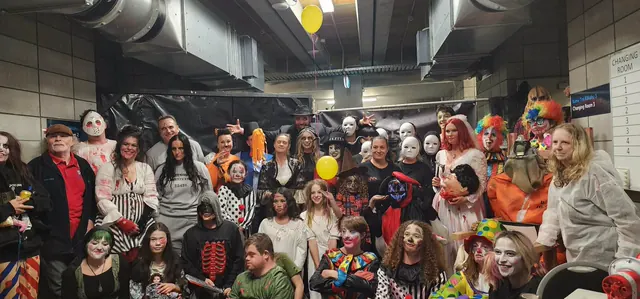 About thirty Northland Horror volunteers crowded together in costume and full makeup backstage at McKay Stadium, clowns, ghouls, masked figures and children in face paint, with balloons drifting above them