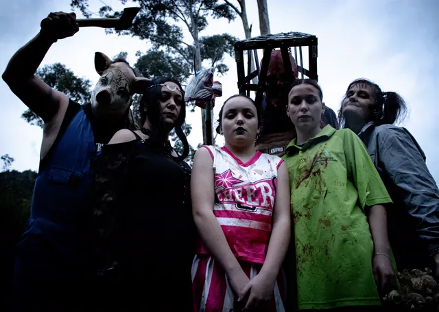 Five Northland Horror performers in costume line up against a pale evening sky - a figure in a pig mask raising an axe, a cheerleader in a blood-spattered uniform, and a scarecrow with a steel cage over its head