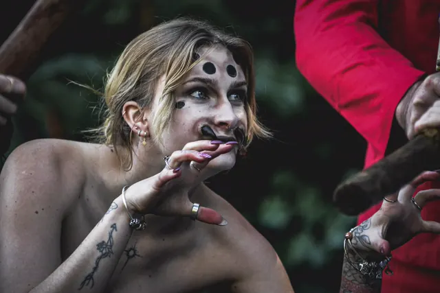 Close-up of the dot-marked character, one clawed hand raised in front of her face, the black mouthpiece curling from either side of her mouth
