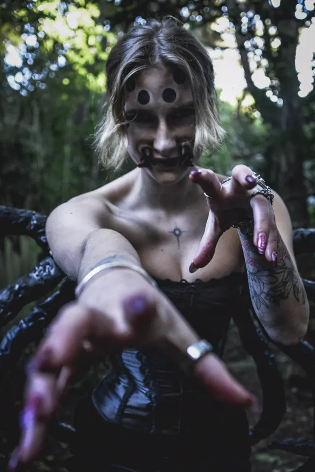 A performer in a black corset, three painted dots across her forehead and a curled black mouthpiece, reaching both hands toward the camera in the bush