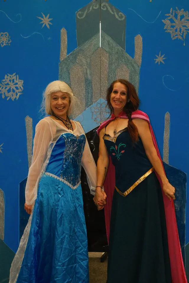 Two volunteers dressed as Elsa and Anna holding hands in front of a hand-painted ice castle backdrop at a family event