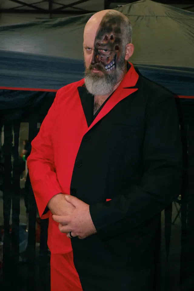 A bearded volunteer in a red and black suit with half his face made up as burned and blistered bone, waiting under a marquee at an event