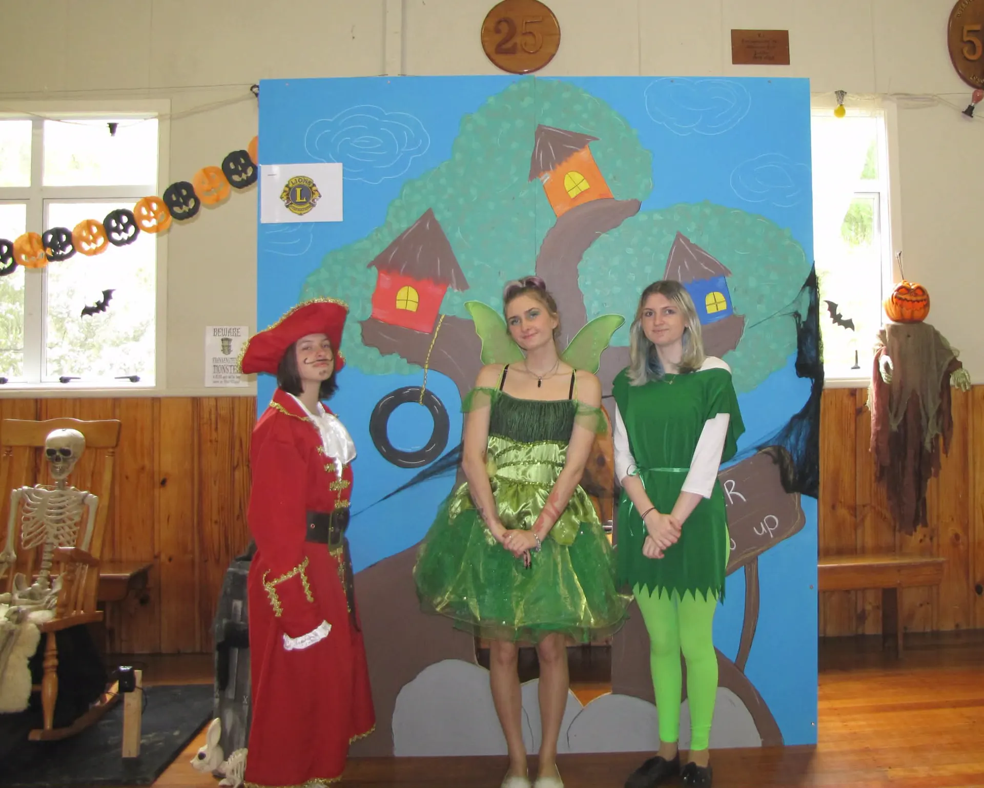 Three volunteers as Captain Hook, Tinker Bell and Peter Pan in front of a painted treehouse backdrop in a community hall strung with paper pumpkins