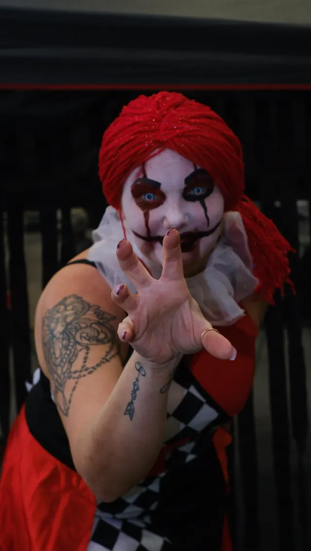 A harlequin in a red yarn wig and black and white diamonds reaching a clawed hand toward the camera at an event