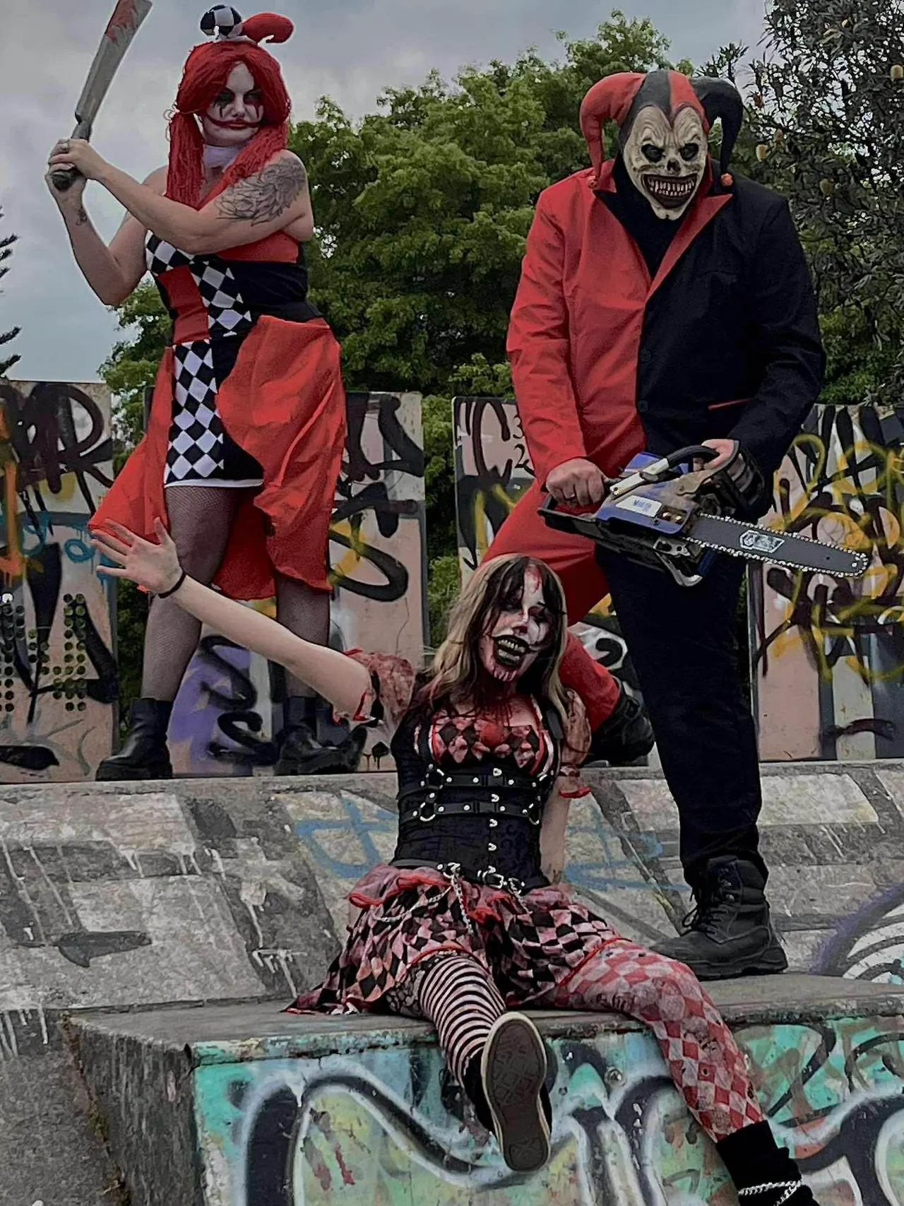 Three harlequin characters staged on a graffiti-covered skate ramp, one raising a machete, one holding a chainsaw and one sprawled between them