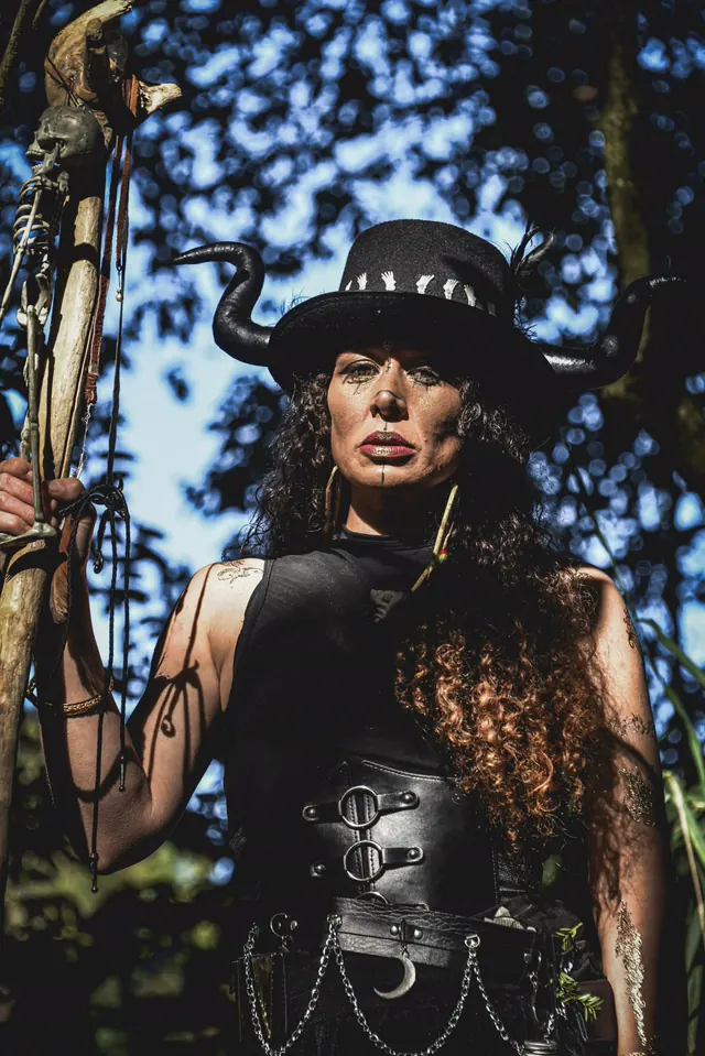 The horned witch photographed from below against a bright canopy, her skeleton-topped staff held beside her