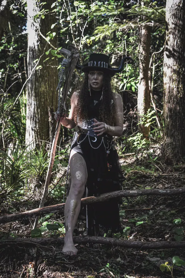 A performer in a horned black top hat and a leather corset holding a bone-topped staff in sunlit bush, gold henna markings up her arms and legs