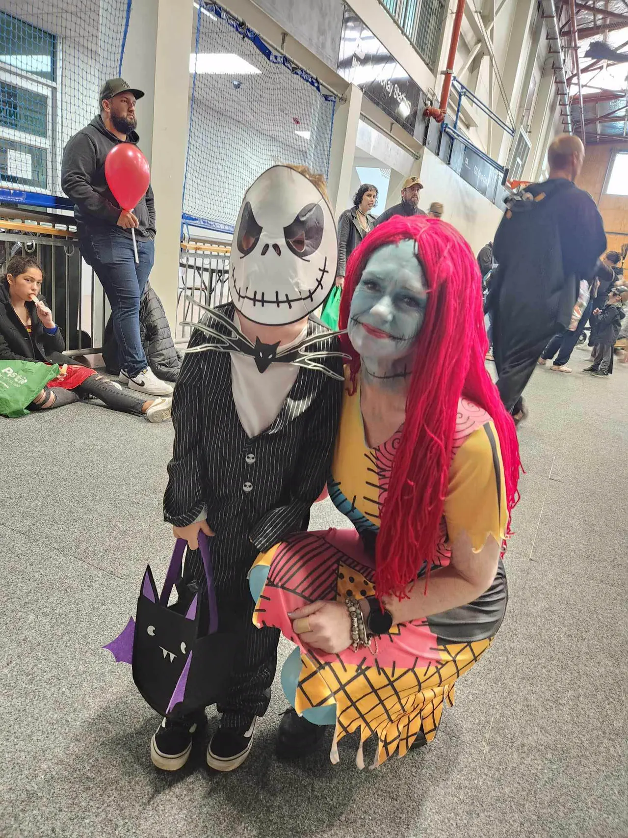 A child in a Jack Skellington mask and pinstripe suit with a bat-shaped trick or treat bag, crouched beside a volunteer made up as Sally, inside McKay Stadium