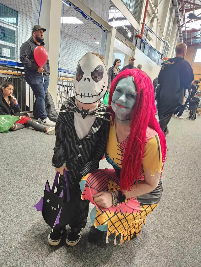 A child in a Jack Skellington mask and pinstripe suit with a bat-shaped trick or treat bag, crouched beside a volunteer made up as Sally, inside McKay Stadium