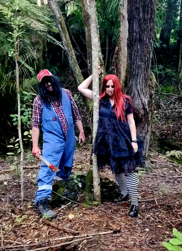 Two characters waiting among the trees, the machete man in blue overalls and a red-wigged doll in a black dress and striped stockings