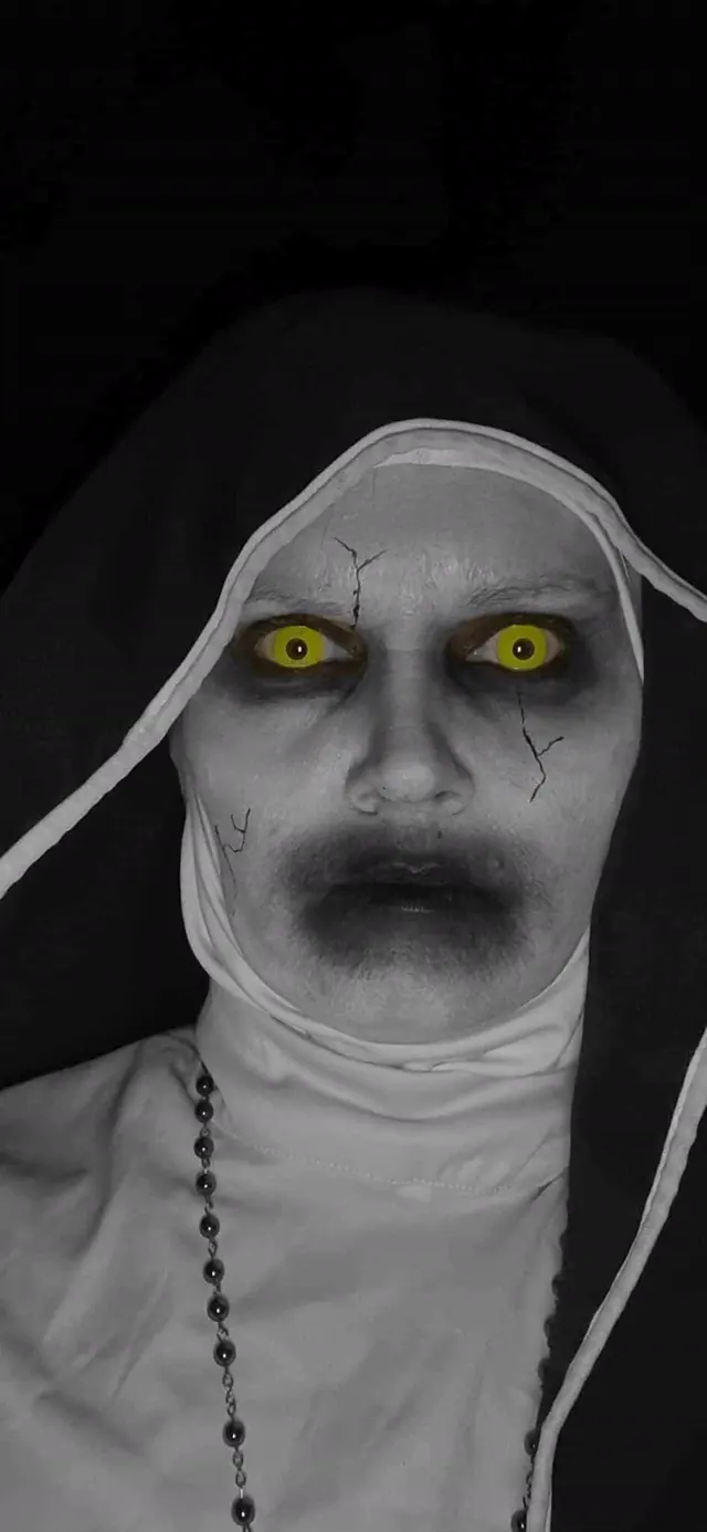 A performer in a nun's habit with cracked grey skin and glowing yellow eyes, lit against near-total black