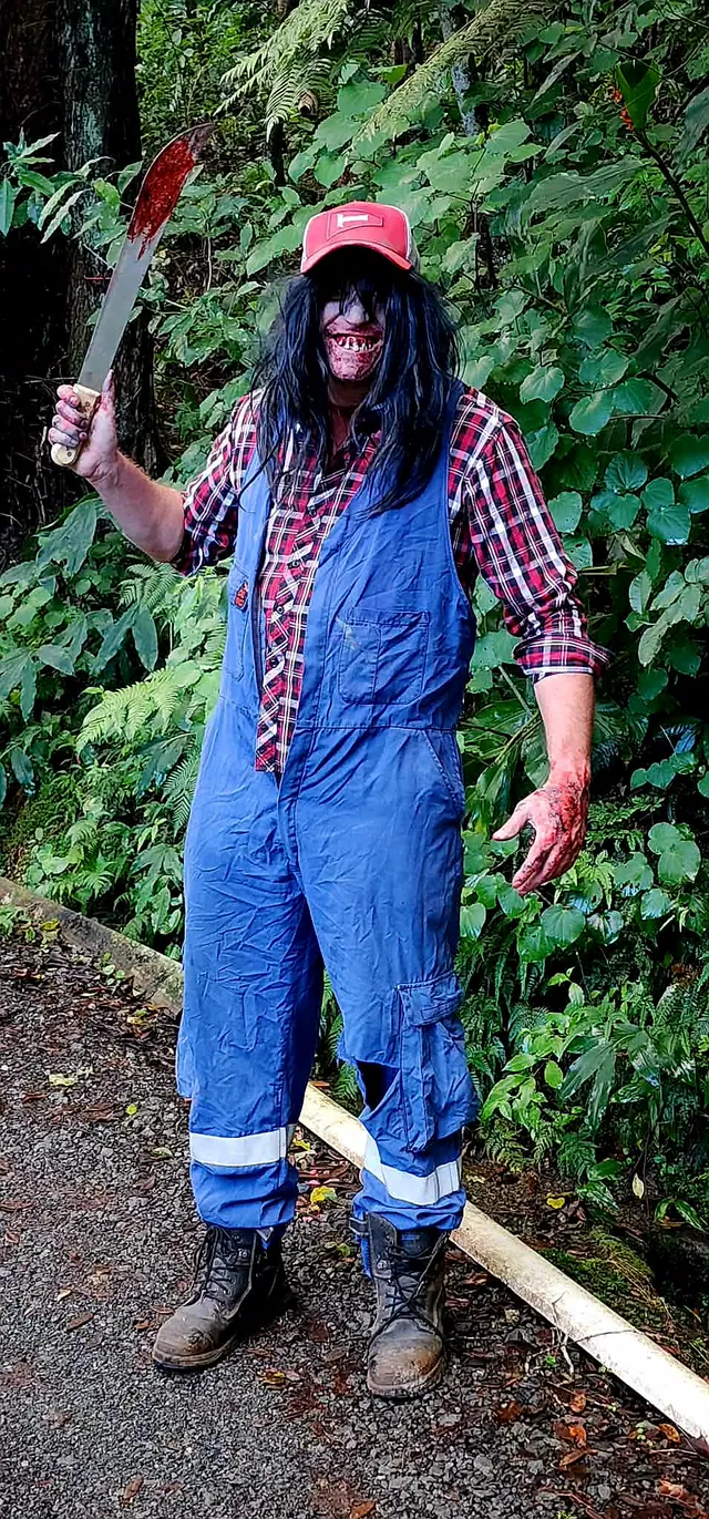 A performer in blue overalls, a red cap and a bloodied mouth prosthetic raising a machete on a bush walking track