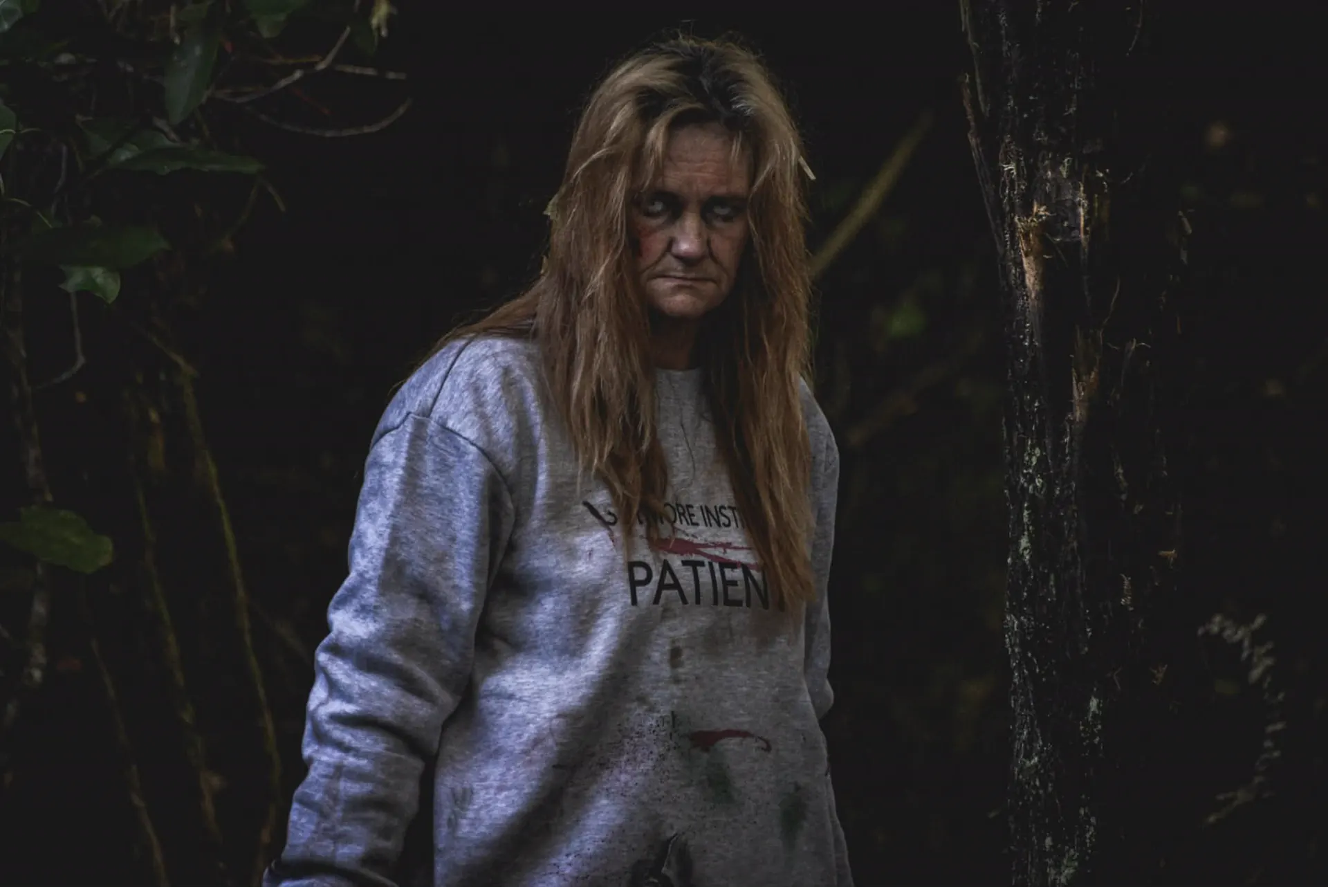 A woman in a blood-stained Slitmore Institution patient sweatshirt stares out of the dark bush, hair matted, a red weal down one cheek