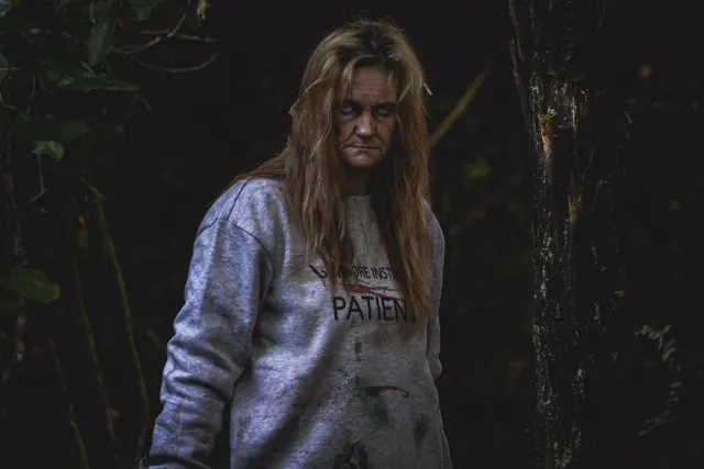 A woman in a blood-stained Slitmore Institution patient sweatshirt stares out of the dark bush, hair matted, a red weal down one cheek