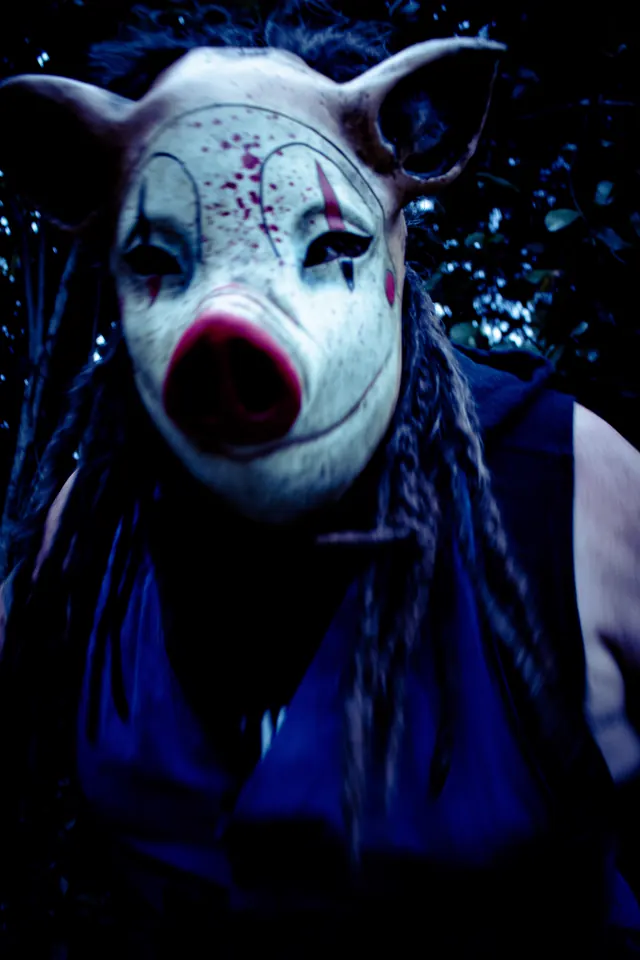Close-up of a blood-spattered pig mask with painted clown markings, framed by long braided hair