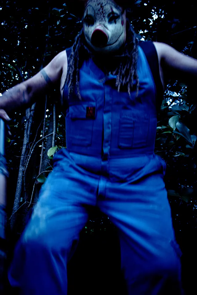 A performer in a bloodied pig mask and blue overalls looms over the camera in the dark bush
