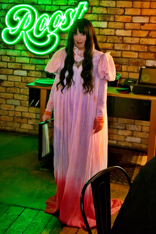 A guest in a long blood-stained prom dress stands under the green neon Roost sign at a Northland Horror quiz night