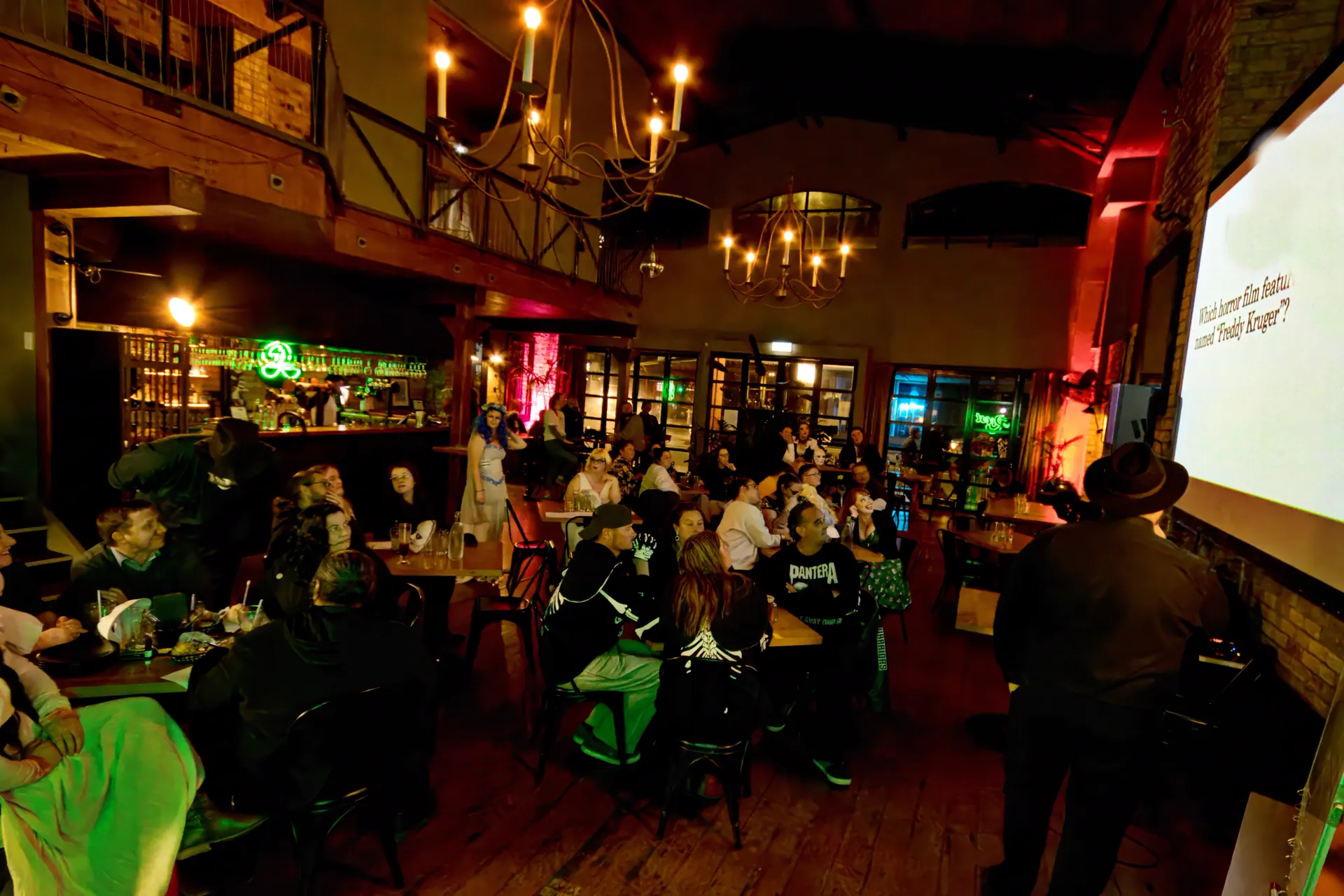 The full quiz night room under chandeliers and red light, every table taken, a question about Freddy Krueger up on the screen
