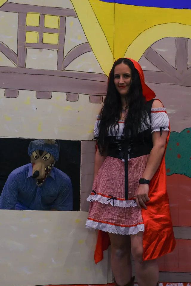A volunteer as Little Red Riding Hood beside a hand-painted cottage set, the wolf in a nightcap leaning out of the window behind her