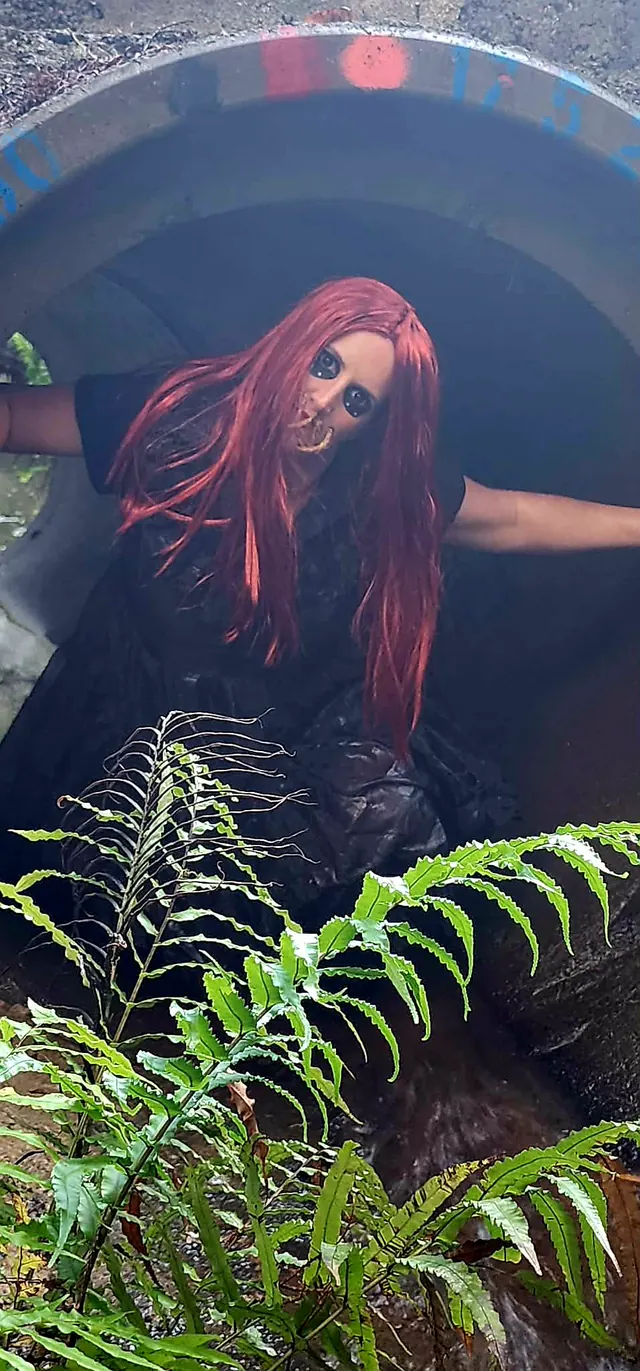 The red-wigged doll crouched inside the mouth of a concrete culvert pipe, framed by ferns