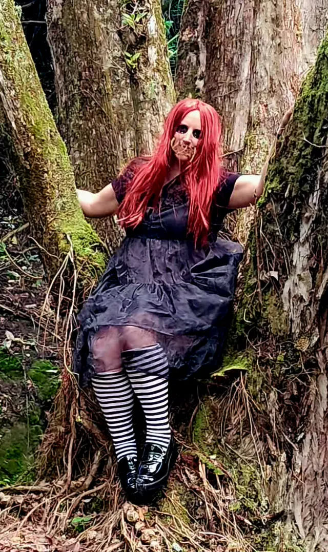 The red-wigged doll character leaning back against a mossy trunk, her eyes blacked out and her mouth stitched shut