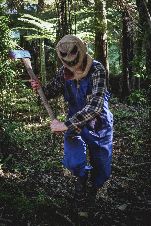 The burlap-hooded scarecrow coming down a fern-lined bush track with a blunt axe held across him