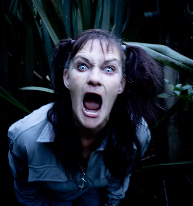 Close-up of a screaming performer with grey skin and pale staring eyes in front of dark flax leaves