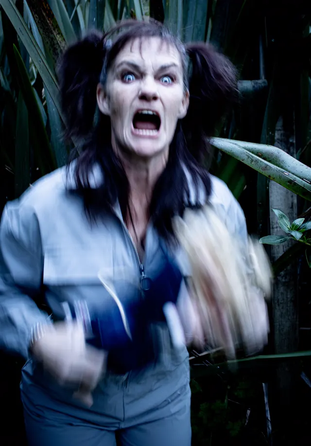 A performer with a corpse-grey face and pigtails screams at the camera as she lunges out of the flax at night