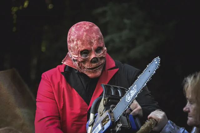 A performer in a raw red skull mask and a red and black suit lifting a chainsaw at dusk