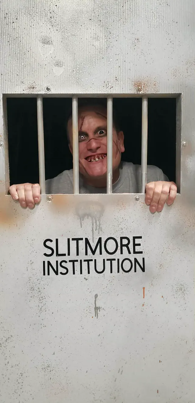 A scarred inmate grinning through the barred hatch of a cell door lettered Slitmore Institution, hands gripping the frame