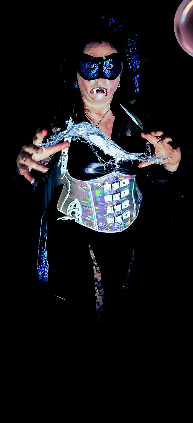 A vampire in a scaled blue masquerade mask and a mirrored corset bares her fangs and reaches for the camera out of near-total darkness