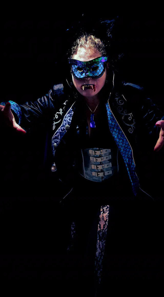 A vampire in a sequinned masquerade mask and an embroidered black jacket spreads her arms in the dark, fangs showing