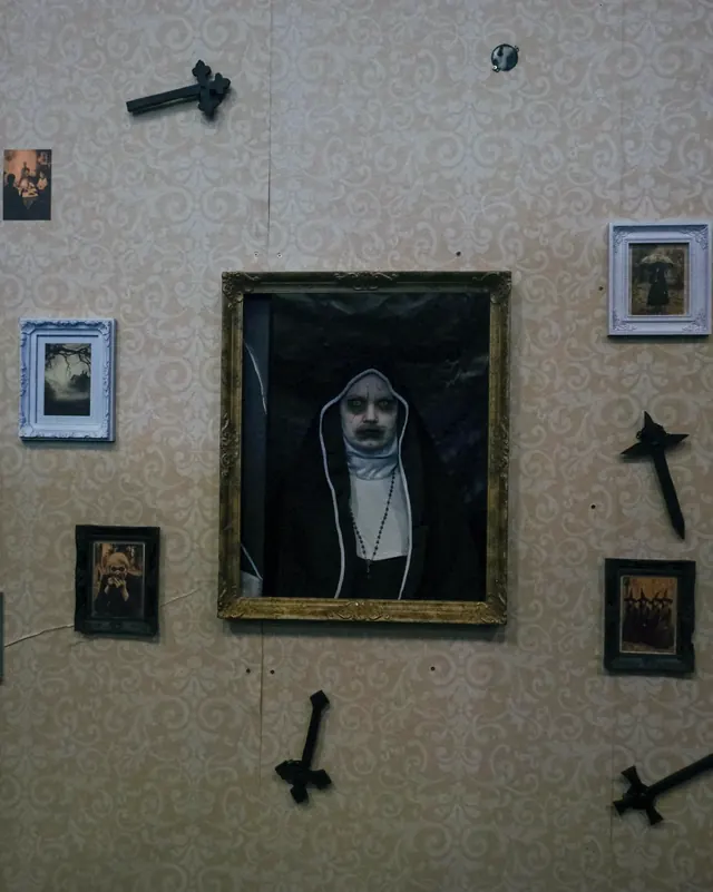 A room built for the walk-through, patterned wallpaper hung with upturned crosses and small framed pictures around a gilt frame with a nun staring out of it