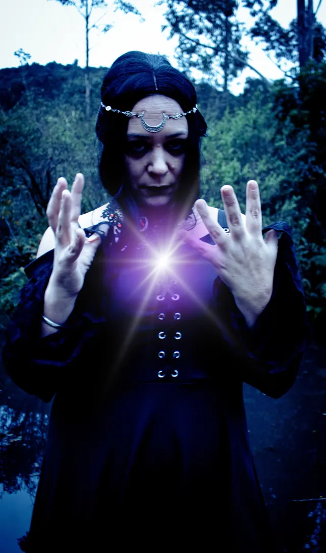 A witch in a black gown and a silver crescent-moon headpiece conjures a burst of violet light between her raised hands beside a bush pond at dusk