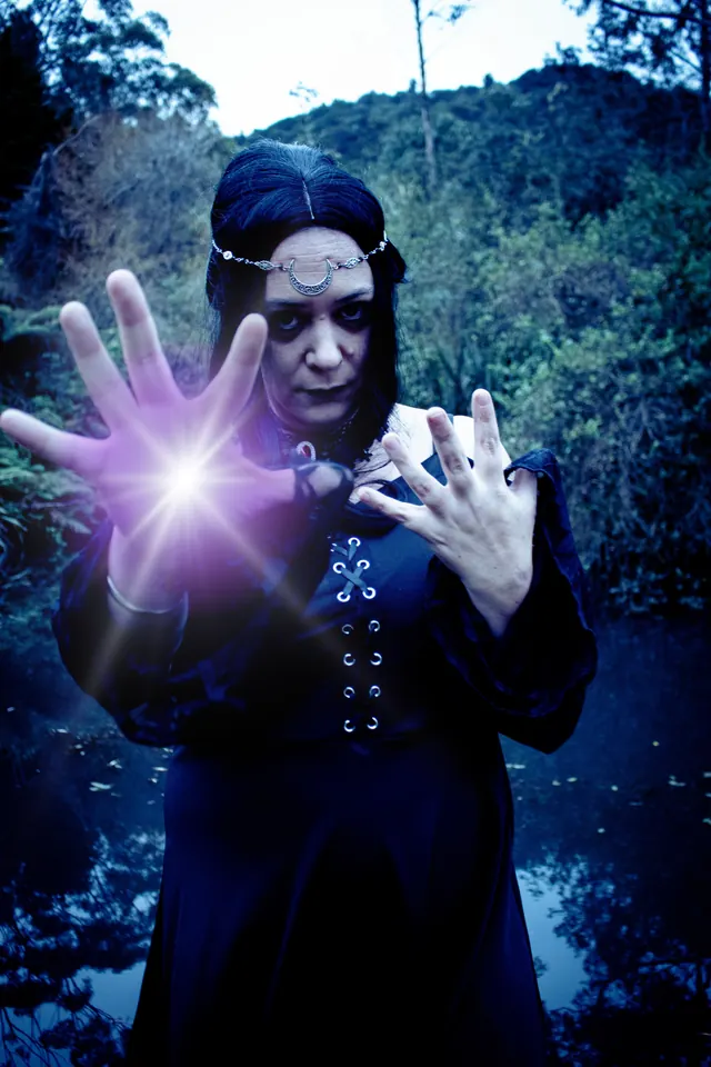 A witch cradles a glowing violet orb in one outstretched hand at the edge of dark bush at dusk